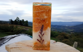 Vela cuadrada naranja de parafina con decoración floral y fondo montañoso